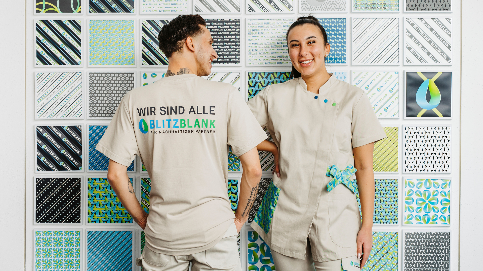 Zwei BLITZBLANK-Mitarbeitende in Arbeitskleidung vor Markenmuster-Wand, Team zeigt Firmenbranding „Wir sind alle BLITZBLANK“